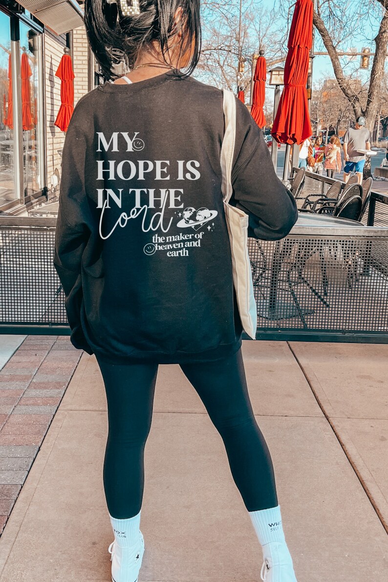 May include: Black sweatshirt with white text reading "MY HOPE IS IN THE Lord" and a graphic of the earth, with the words "the maker of heaven and earth." The sweatshirt is worn with black leggings and white trainers.