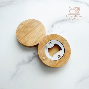 May include: A wooden bottle opener with a round, flat top and a circular opening for opening bottles. The opener is made of light brown wood and has a silver metal bottle opener insert. The opener is on a white marble surface.