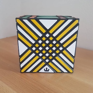 Könnte beinhalten: Ein mehrfarbiger Puzzle-Würfel mit einem komplexen geometrischen Muster. Der Würfel hat schwarze, weiße und gelbe diagonale Linien mit einem kleinen Kronenlogo auf einer Seite. Der Würfel steht auf einer Holzoberfläche.