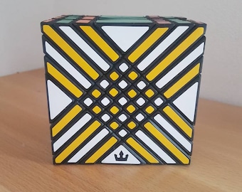 May include: A multi-colored puzzle cube with a complex geometric pattern. The cube features black, white, and yellow diagonal lines, with a small crown logo on one side. The cube is sitting on a wooden surface.