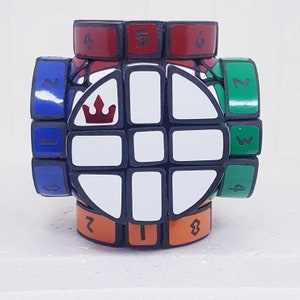 Puede incluir: Un cubo de rompecabezas blanco y negro con un diseño de corona en un lado. El cubo tiene los números del 1 al 6 en los lados. El cubo está hecho de plástico y tiene un acabado brillante.