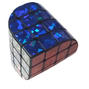 Puede incluir: Un cubo de rompecabezas tridimensional curvo con una cuadrícula de cuadrados pequeños. La superficie superior muestra un acabado holográfico azul profundo y reflectante, mientras que los paneles laterales presentan una superficie metálica plateada.