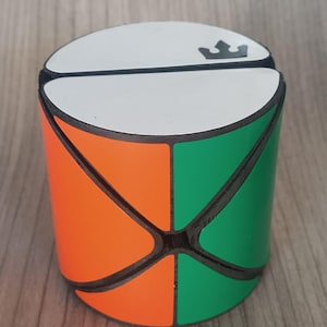 Könnte beinhalten: Ein zylindrisches Puzzlespielzeug mit weißer Oberseite und einem Kronenlogo. Die Seiten sind in orangefarbene und grüne Abschnitte unterteilt, die durch schwarze Linien getrennt sind. Das Puzzle ist zum Drehen und Wenden konzipiert.