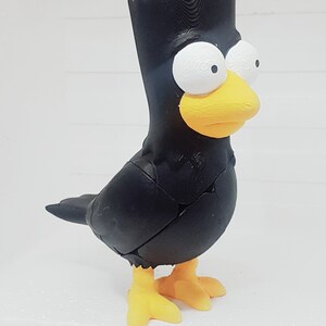 May include: A black bird-shaped ornament with a yellow beak and feet, and white eyes. The figurine has a distinctive hairstyle and is standing on a white surface. The bird is a unique decorative item.