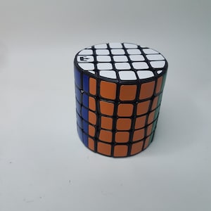 Puede incluir: Un juguete de rompecabezas cilíndrico con una parte superior blanca y negra, lados naranjas y detalles azules y verdes. El rompecabezas tiene un patrón de cuadrícula y la letra "E" es visible en la parte superior. El juguete está diseñado para girar y resolver el rompecabezas.