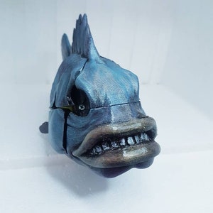 May include: A blue and gray 3D printed fish sculpture with a toothy grin and a spiky fin. The fish has a detailed, textured surface and a realistic appearance.