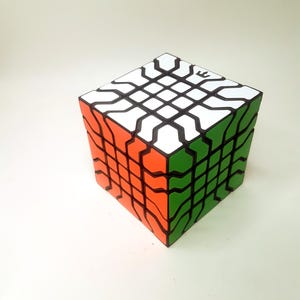 Può includere: Un cubo di puzzle 3x3x3 nero, bianco, arancione e verde con un motivo unico su ogni lato. Il cubo è fatto di plastica e ha una finitura liscia e lucida.