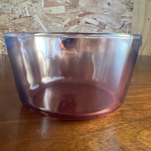 May include: A clear, amethyst-coloured glass bowl with a slightly flared shape. The bowl sits on a wooden surface, reflecting the light. The bowl's smooth surface and deep colour suggest it could be used for serving or display.
