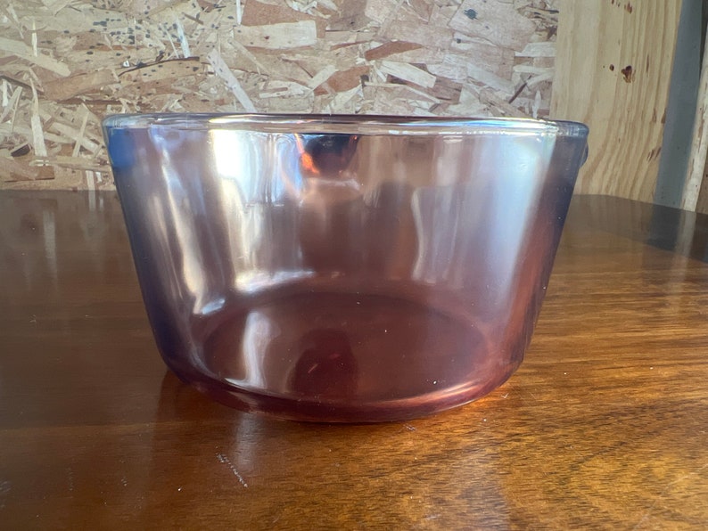 May include: A clear, amethyst-coloured glass bowl with a slightly flared shape. The bowl sits on a wooden surface, reflecting the light. The bowl's smooth surface and deep colour suggest it could be used for serving or display.