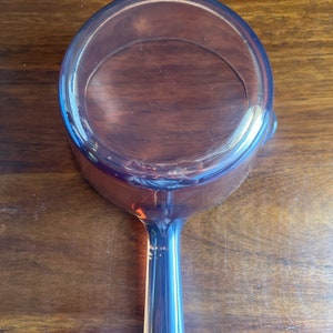 May include: An amber-coloured glass saucepan with a long handle. The pan is round with a slightly flared rim and a smooth interior. The handle is solid and tapers towards the end. The saucepan is sitting on a wooden surface.
