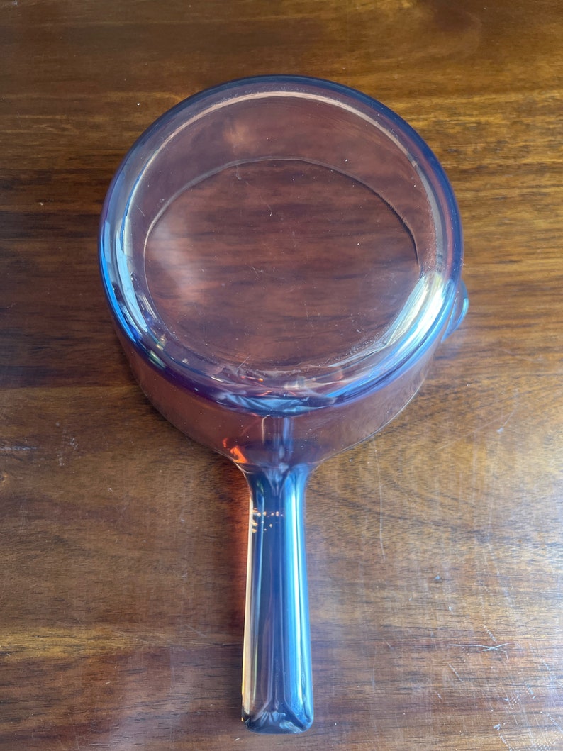 May include: An amber-coloured glass saucepan with a long handle. The pan is round with a slightly flared rim and a smooth interior. The handle is solid and tapers towards the end. The saucepan is sitting on a wooden surface.