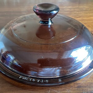 May include: A brown glass lid with a rounded shape and a dark brown knob. The lid is transparent, allowing a view of the contents below. The text "VINTAGE V-1-B" is printed on the rim. This lid is likely for a casserole dish.