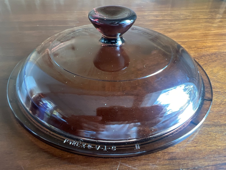 May include: A brown glass lid with a rounded shape and a dark brown knob. The lid is transparent, allowing a view of the contents below. The text "VINTAGE V-1-B" is printed on the rim. This lid is likely for a casserole dish.