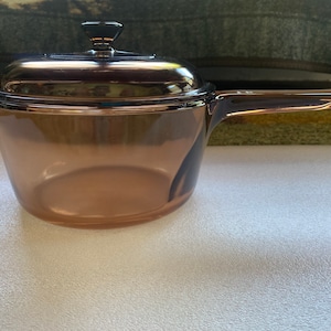 May include: An amber-tinted glass saucepan with a lid. The saucepan features a long handle and is designed for use on a hob. The lid has a small, black knob. The cookware is made of glass.