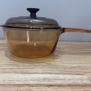 May include: A clear glass saucepan with a brown lid and a handle. The saucepan is sitting on a wooden surface.