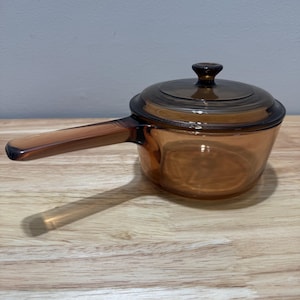 Puede incluir: Una cacerola de vidrio ámbar vintage con tapa y asa a juego. La cacerola está sobre una superficie de madera. El asa está unida al lateral de la cacerola. La tapa tiene un pequeño pomo en la parte superior.