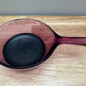 Può includere: Una padella vintage in vetro trasparente color rubino con un inserto rotondo nero. La padella ha un lungo manico e una forma a ciotola arrotondata. La padella è appoggiata su una superficie di legno.