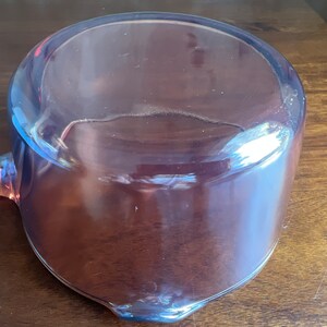 May include: A clear, pink-tinted glass ladle with a rounded bowl and a long handle. The ladle is made of transparent glass, allowing light to pass through. The handle extends from the side of the bowl.
