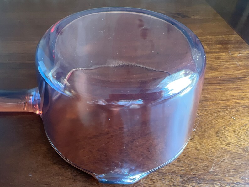 May include: A clear, pink-tinted glass ladle with a rounded bowl and a long handle. The ladle is made of transparent glass, allowing light to pass through. The handle extends from the side of the bowl.