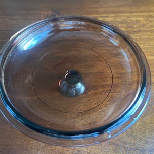 May include: A transparent glass lid with a dark brown knob in the centre. The lid is circular with a slightly raised edge and a circular design. The knob is cone-shaped and appears to be made of the same glass material.