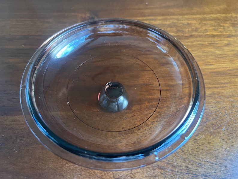 May include: A transparent glass lid with a dark brown knob in the centre. The lid is circular with a slightly raised edge and a circular design. The knob is cone-shaped and appears to be made of the same glass material.
