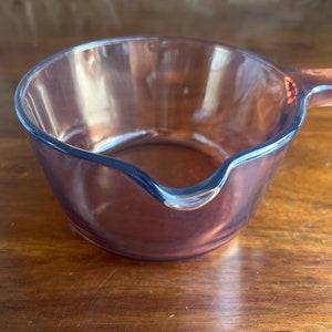 May include: A clear, amber-coloured glass saucepan with a pouring spout and a handle. The small pan is made of transparent glass, allowing visibility of the contents. The handle is also amber-coloured. The pan sits on a wooden surface.