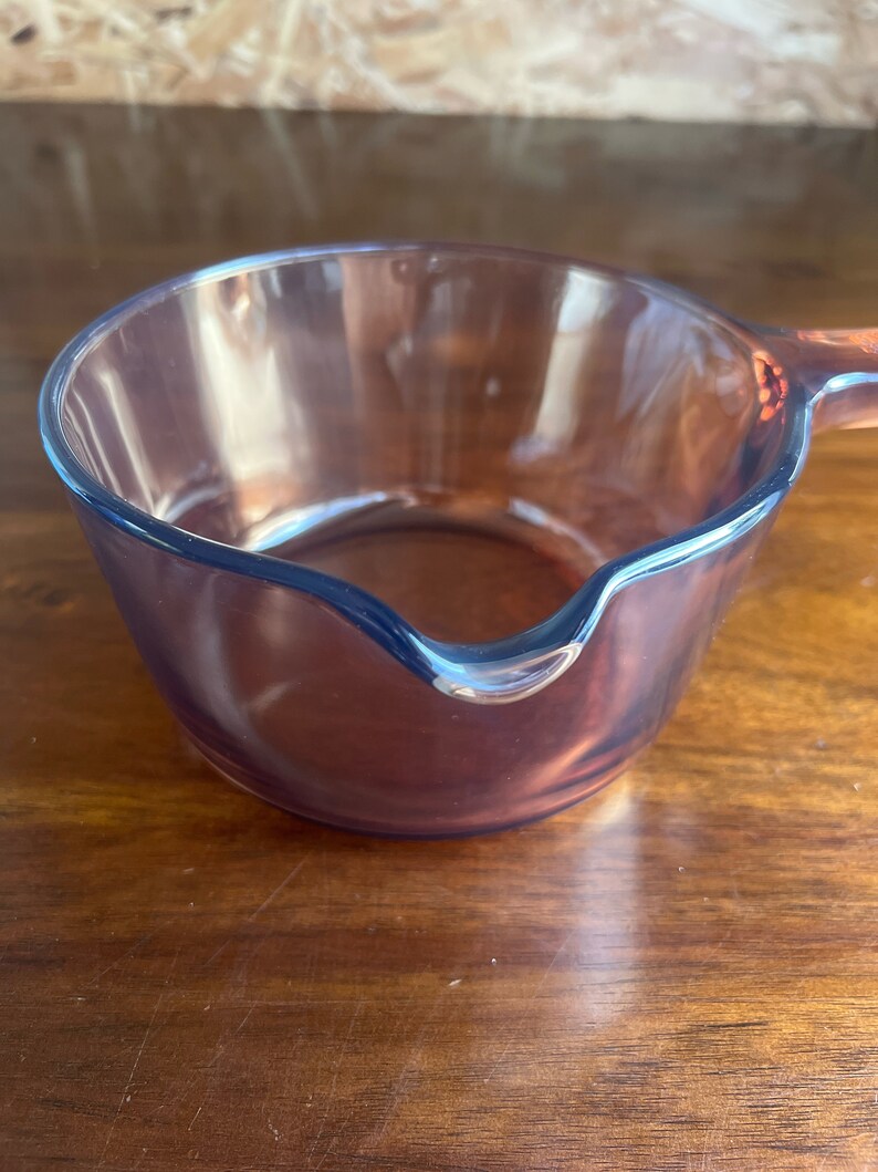 May include: A clear, amber-coloured glass saucepan with a pouring spout and a handle. The small pan is made of transparent glass, allowing visibility of the contents. The handle is also amber-coloured. The pan sits on a wooden surface.