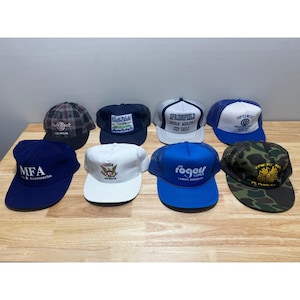 Puede incluir: Una colección de ocho gorras de béisbol en varios colores y diseños. Las gorras presentan logotipos y texto, incluyendo "Hard Rock", "Springfield" y "Jim Roger Lures". Los estilos incluyen respaldos de malla, patrones de cuadros y colores sólidos. Las gorras se exhiben sobre una superficie de madera clara.