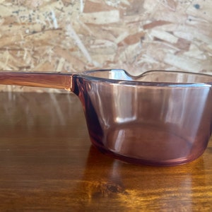 May include: A clear, vintage-style glass saucepan with a long handle. The saucepan has a rounded bowl shape and a handle that extends from the side. The glass has a translucent, purple-brown hue. The handle is a solid, darker shade of the same colour.