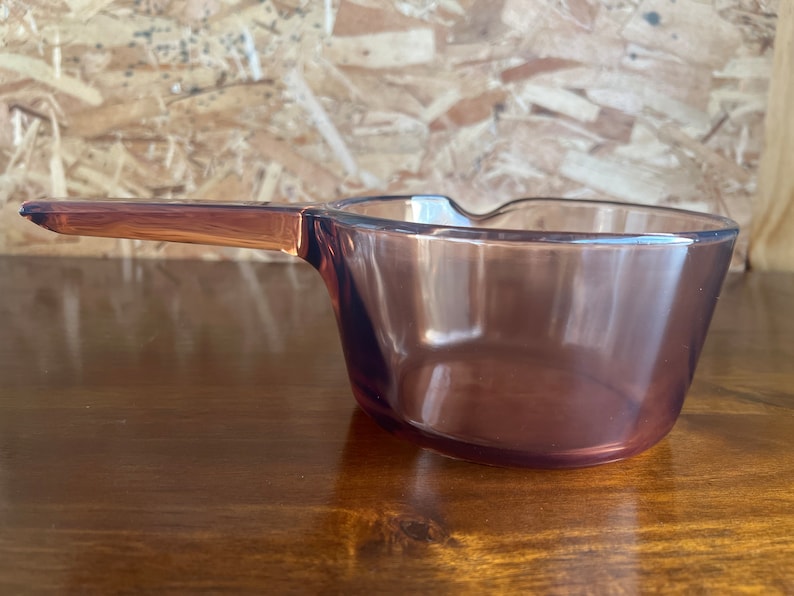 May include: A clear, vintage-style glass saucepan with a long handle. The saucepan has a rounded bowl shape and a handle that extends from the side. The glass has a translucent, purple-brown hue. The handle is a solid, darker shade of the same colour.