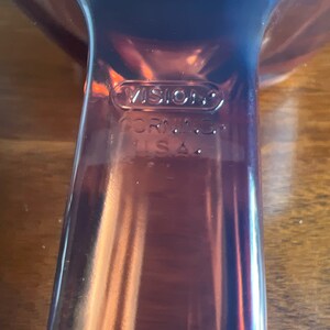 May include: Close-up of a brown Vision Corning USA glass serving dish. The dish has a handle and a curved design. The word "VISION" is embossed on the glass, along with "CORNING" and "U.S.A."