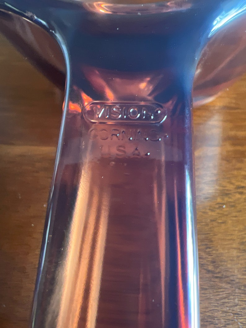 May include: Close-up of a brown Vision Corning USA glass serving dish. The dish has a handle and a curved design. The word "VISION" is embossed on the glass, along with "CORNING" and "U.S.A."