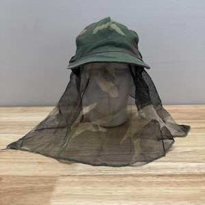 May include: A camouflage hat with a built-in mosquito net. The hat features a green and brown camouflage pattern on the crown and brim. The net is black and drapes down, providing protection from insects. The hat is designed for outdoor activities.