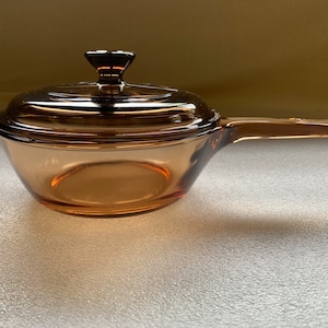 Può includere: Una casseruola vintage in vetro color ambra con coperchio. La casseruola ha un manico ed è progettata per la cottura sul piano cottura. Il coperchio ha un piccolo pomello per una facile manipolazione. Questo utensile da cucina è un classico.