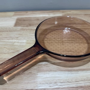 Puede incluir: Una sartén de vidrio transparente de color ámbar con un mango largo. La sartén tiene una superficie de cocción redondeada y un borde negro. El mango también es de color ámbar y tiene una forma suave y redondeada. La sartén está sobre una superficie de madera.