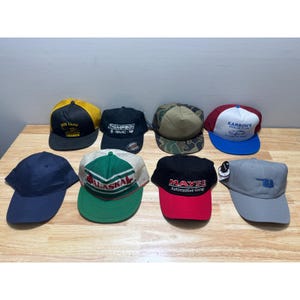 Könnte beinhalten: Eine Sammlung von acht Baseballkappen in verschiedenen Farben und Designs. Die Kappen zeigen Logos und Texte, darunter "Red Lane Canada", "Thompson GMC" und "Alaska". Einige Kappen haben Mesh-Rücken und kontrastierende Krempen.