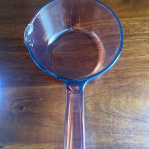 May include: A clear glass saucepan with a handle and a pouring spout. The saucepan is made of transparent glass and has a round shape. The handle is rectangular and has the brand name "VISION" printed on it.