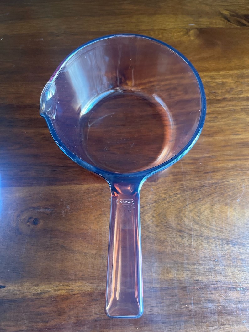 May include: A clear glass saucepan with a handle and a pouring spout. The saucepan is made of transparent glass and has a round shape. The handle is rectangular and has the brand name "VISION" printed on it.