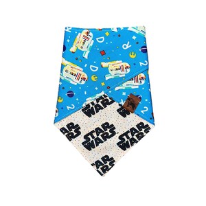May include: A blue and white reversible dog bandana with a Star Wars theme. One side features a repeating pattern of R2-D2 robots, planets, and the number 2. The other side features a repeating pattern of the words "Star Wars" in black on a white background.
