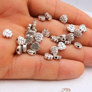 May include: A pile of silver leaf-shaped beads with a hole in the center of each bead. The beads are scattered on a hand.
