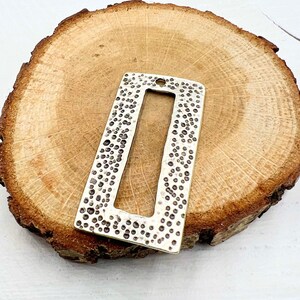 May include: A silver-toned metal rectangle with a textured surface and a rectangular cutout in the center. The rectangle is on a wooden surface.