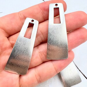 May include: Two silver-toned metal earring blanks with a rectangular cut-out in the center. The blanks are slightly curved and have a textured finish.