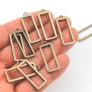 Könnte beinhalten: Nahaufnahme einer Hand, die eine Gruppe von rechteckigen Anhänger-Rahmen aus antikem Goldmetall hält. Die Rahmen sind offen und haben eine glatte, leicht strukturierte Oberfläche.