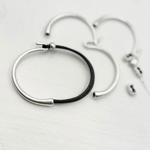 May include: A silver bracelet with a black leather band. The bracelet is open and has a simple design.