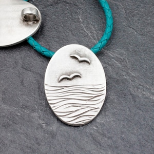 May include: Silver pendant necklace with a turquoise leather cord. The pendant features a detailed design of two birds flying over a wavy ocean.