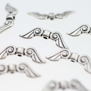 May include: Silver-toned metal angel wing charms with a decorative swirl design. The charms have a hole in the center for attaching to jewelry or other crafts.
