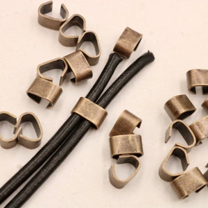 May include: A close-up of antique gold metal cord ends for leather cord. The cord ends are designed to hold two cords together. There are multiple cord ends scattered around the image.