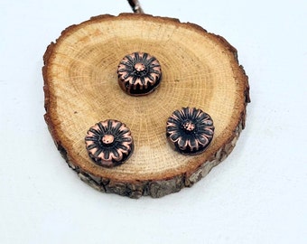 May include: Three copper-colored metal flower-shaped embellishments on a wooden surface.
