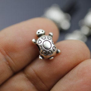 May include: A silver turtle charm with a detailed shell design. The charm is small and has a hole for attaching to a bracelet or necklace.