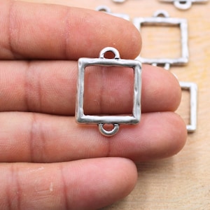 Könnte beinhalten: Silberfarbener quadratischer Metallrahmen mit zwei Schlaufen oben. Der Rahmen hat eine strukturierte Oberfläche und ist für die Schmuckherstellung gedacht.